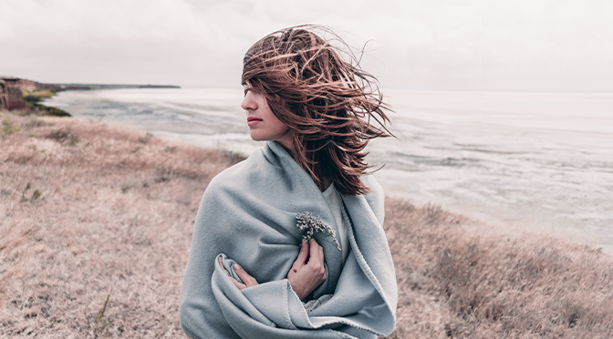 Attraktive junge Frau, die auf einem windigen kalten Strand steht, umhüllt von warmer Decke mit Blume in der Hand.