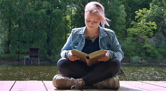 junge Frau mit Buch in der Hand sitzt auf einem Steg am Wasser