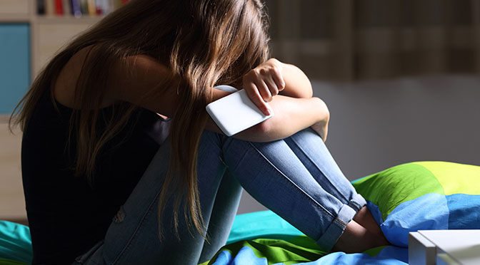 Einzelne traurige Teenager, die ein Mobiltelefon halten, das auf dem Bett sitzt, in ihrem Schlafzimmer mit dunklem Licht auf dem Hintergrund