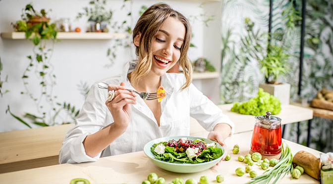 Junge Frau, die gesundes Essen isst, sitzt im schönen Innern mit grünen Blumen auf dem Hintergrund