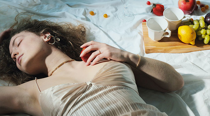 Frau liegt mit geschlossenen Augen auf dem Bett, einen Pfirisch in der Hand
