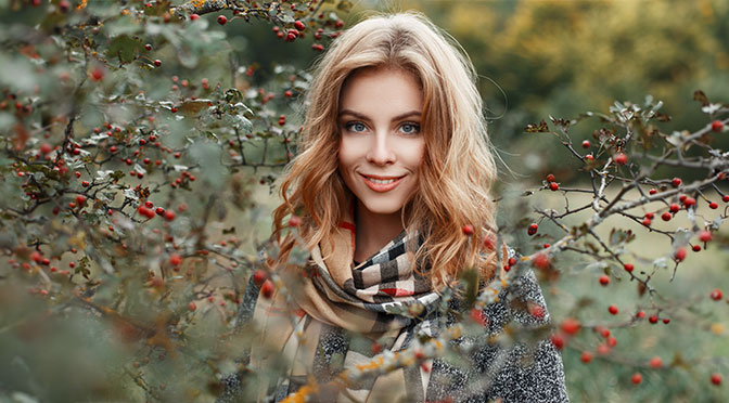 Reine junge Frau mit blondem Haar mit blauen Augen mit einem schönen Lächeln in einem stilvollen warmen Mantel in einem vintage Schal aus Schachbrettmuster steht im Wald zwischen den trockenen Ästen der Bersche Asche. Fröhliches Mädchen.