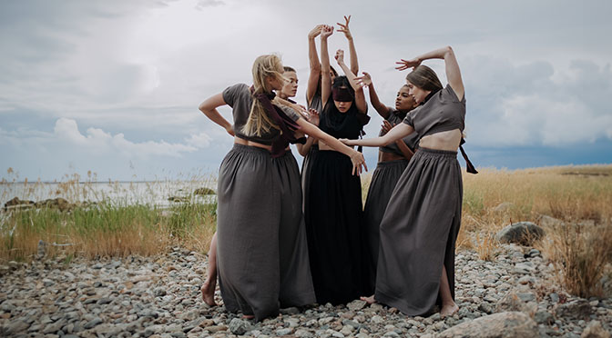 Frauen tanzen am Ufer eines Sees, eine von ihnen mit verbundenen Augen