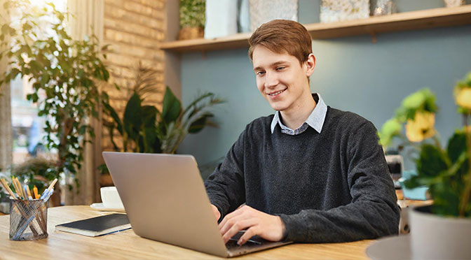 Ein junger Mann Anfang zwanzig sitzt an einem Schreibtisch und tippt etwas in seinen Laptop. Er lächelt zufrieden