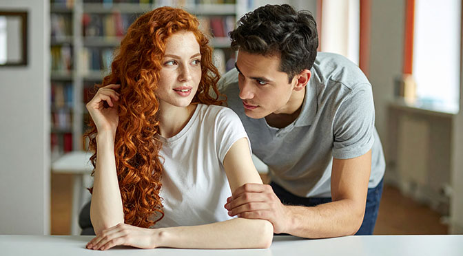 Clara, a woman in her mid-twenties, leans against the table in a library. She is slim with soft curves, wearing a t-shirt. Her red hair falls down in curls. Elias, a young man with sharp features, steps closer. He touches her arm.