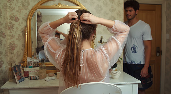 A woman, approximately 25 years old, sits at her dressing table, wearing a see-through nightgown. She has long hair, which she is tying into a ponytail with both hands. Her boyfriend stands a little way off, watching her.