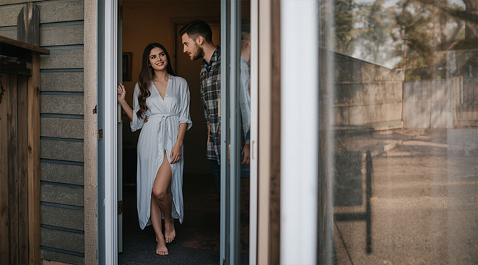 A young woman, 23 years old, with long brown hair and a gentle smile, stands barefoot in the doorway, wearing a soft white dressing gown that drapes elegantly around her slender figure, as she opens the front door to reveal a man of similar age, with short dark hair and warm brown eyes, who gazes at her with affection and adoration, his facial expression conveying a deep sense of love and connection, with a soft focus on the couple to emphasize the intimacy of the moment, set against a warm and inviting background with a mix of earthy tones and soft natural light pouring in from the outside.