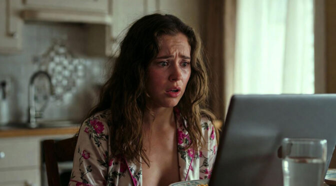 A woman sits at the kitchen table in her pajamas, staring in horror at the open laptop in front of her.