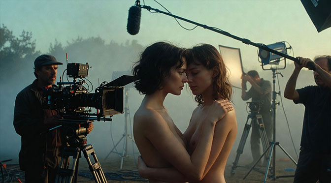 Two women with bare upper bodies, tightly entwined in an intimate embrace. Surrounding them: a man operating a professional movie camera, lighting technicians with equipment, and a man holding a large microphone boom, film set atmosphere