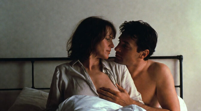 Eine 45-jährige Frau sitzt auf ihrem Bett und trägt eine leichte Bluse. Neben ihr ein etwas jüngerer Mann, der zärtlich mit seiner Hand unter ihre Bluse fasst. Beide schauen sich sehr verliebt in die Augen.